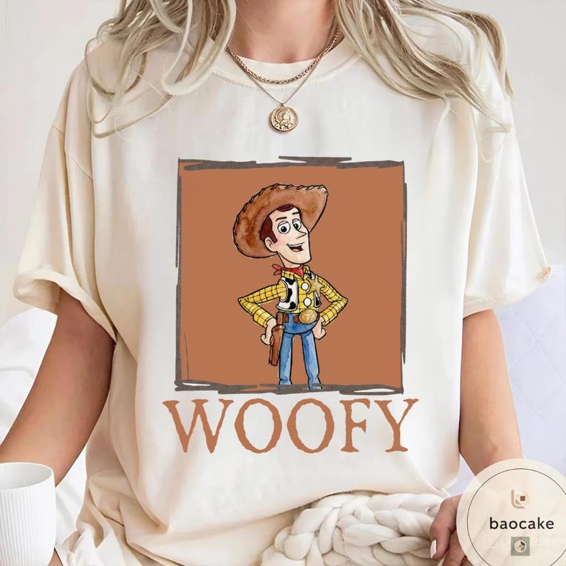 K&ouml;nnte beinhalten: Ein cremefarbenes Kurzarm-T-Shirt mit einer Grafik eines Cartoon-Cowboy-Charakters, der einen braunen Hut, ein gelbes kariertes Hemd und eine blaue Hose tr&auml;gt, in einem braunen quadratischen Rahmen, mit dem Wort WOOFY in gro&szlig;en braunen Buchstaben.