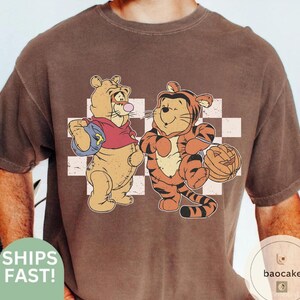 May include: Brown t-shirt featuring a cartoon illustration of two characters, one dressed as a bear and the other as a tiger, standing in front of a white and light brown checkered background. The bear is wearing a red shirt and carrying a blue backpack. The tiger is holding a carved pumpkin.