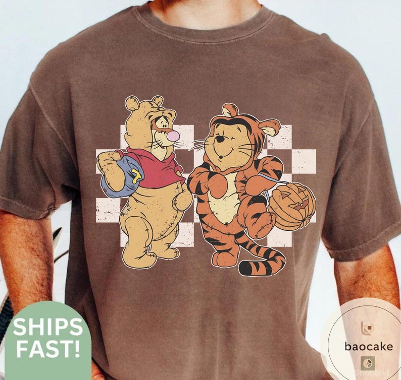 May include: Brown t-shirt featuring a cartoon illustration of two characters, one dressed as a bear and the other as a tiger, standing in front of a white and light brown checkered background. The bear is wearing a red shirt and carrying a blue backpack. The tiger is holding a carved pumpkin.