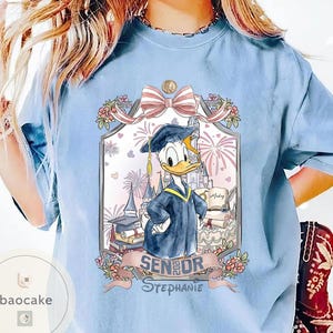 May include: Light blue t-shirt featuring a graduation-themed graphic with Donald Duck in a cap and gown. The design includes a castle, fireworks, and the text "Senior 2023 Stephanie."
