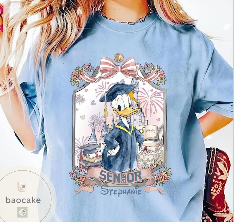 May include: Light blue t-shirt featuring a graduation-themed graphic with Donald Duck in a cap and gown. The design includes a castle, fireworks, and the text "Senior 2023 Stephanie."