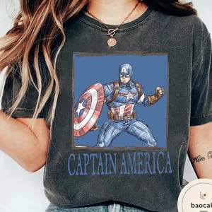 May include: Dark gray t-shirt featuring a Captain America graphic. The design shows Captain America in a blue and red costume with his shield, set against a blue background. The words "CAPTAIN AMERICA" are printed below the image.