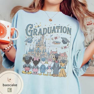 May include: Light blue t-shirt with the word "Graduation" in a vintage font, featuring a Disney castle and cartoon characters in graduation attire. The shirt includes a coffee mug with a sun design.