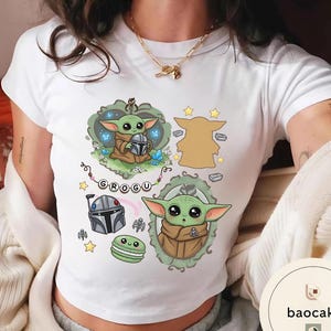 May include: A white short-sleeved t-shirt featuring a graphic print of the character Grogu. The design includes two illustrations of the green creature, a silver helmet, a green macaron, yellow stars, and a banner with the text GROGU.