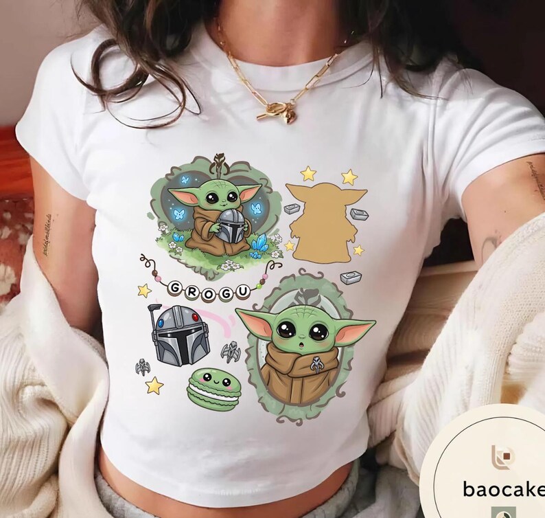 May include: A white short-sleeved t-shirt featuring a graphic print of the character Grogu. The design includes two illustrations of the green creature, a silver helmet, a green macaron, yellow stars, and a banner with the text GROGU.