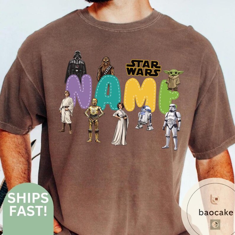 May include: Brown t-shirt featuring Star Wars characters and the word "NAME" in colorful bubble letters. Characters include Darth Vader, Chewbacca, Luke Skywalker, C-3PO, Princess Leia, R2-D2, a Stormtrooper, and Grogu.