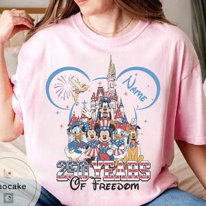 Peut inclure: T-shirt rose clair avec un motif color&eacute; de personnages Disney en tenue patriotique. Le motif comprend un ch&acirc;teau, des feux d'artifice et le texte "250 YEARS OF FREEDOM" avec un espace pour un nom. Le t-shirt a un col rond.