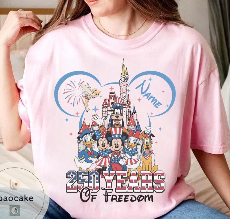 Peut inclure: T-shirt rose clair avec un motif color&eacute; de personnages Disney en tenue patriotique. Le motif comprend un ch&acirc;teau, des feux d'artifice et le texte "250 YEARS OF FREEDOM" avec un espace pour un nom. Le t-shirt a un col rond.