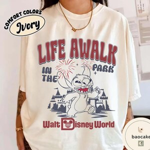 May include: Ivory-colored t-shirt with a graphic featuring Stitch from Disney's Lilo & Stitch. The design includes the text "LIFE AWALK IN THE PARK" and "Walt Disney World" in red and blue. The shirt has a Comfort Colors label.