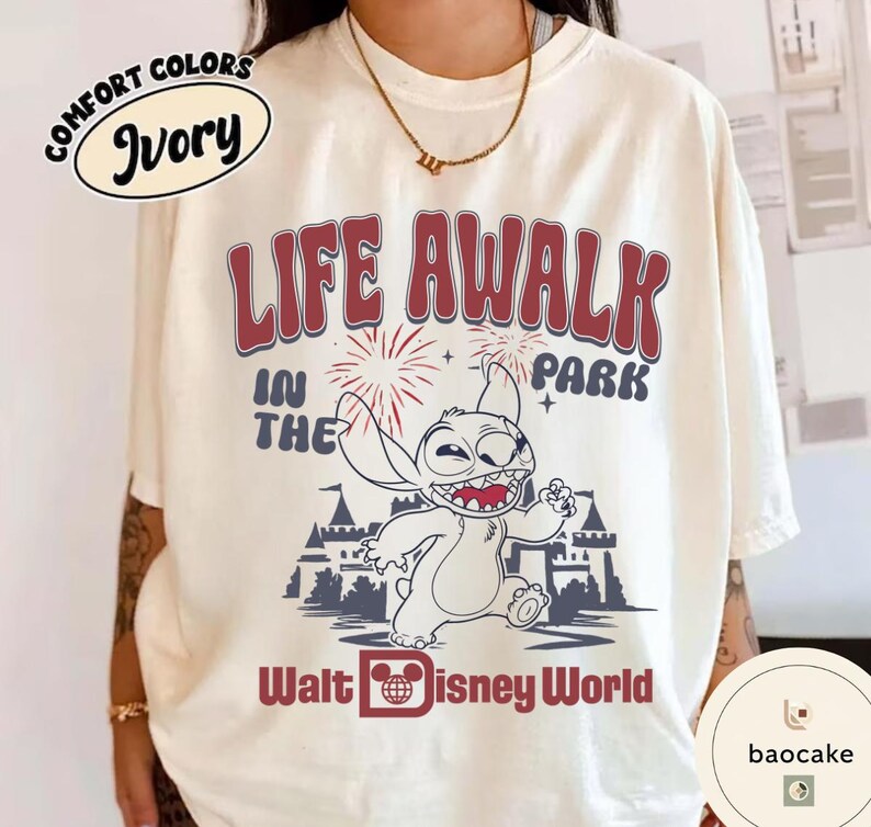 May include: Ivory-colored t-shirt with a graphic featuring Stitch from Disney's Lilo & Stitch. The design includes the text "LIFE AWALK IN THE PARK" and "Walt Disney World" in red and blue. The shirt has a Comfort Colors label.