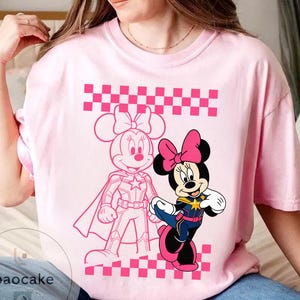 May include: Pink t-shirt featuring a graphic of Minnie Mouse in two different styles. One is a pink outline, and the other is a full-color image of Minnie Mouse in a superhero outfit. The shirt has a pink and white checkered pattern.