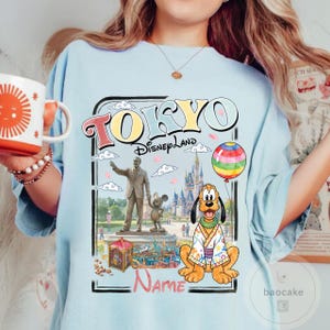 Puede incluir: Camiseta azul claro con un gr&aacute;fico colorido de Tokyo Disneyland. El dise&ntilde;o incluye las palabras "TOKYO Disneyland" en una fuente retro, una estatua, Pluto con un kimono y un globo de arco&iacute;ris. Hay espacio para un nombre.
