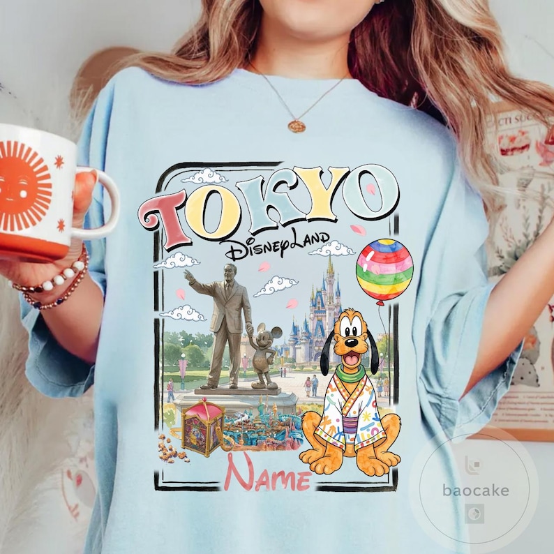 Puede incluir: Camiseta azul claro con un gr&aacute;fico colorido de Tokyo Disneyland. El dise&ntilde;o incluye las palabras "TOKYO Disneyland" en una fuente retro, una estatua, Pluto con un kimono y un globo de arco&iacute;ris. Hay espacio para un nombre.