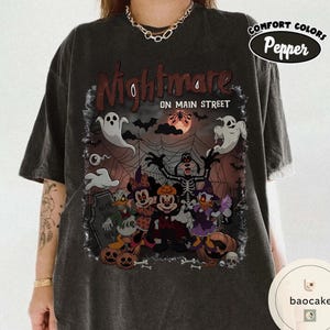May include: Dark gray t-shirt featuring a Halloween-themed graphic with the text "Nightmare on Main Street." The design includes cartoon characters dressed in Halloween costumes, ghosts, pumpkins, and a full moon. The shirt has a Comfort Colors Pepper label.