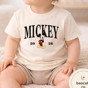 May include: Cream-colored t-shirt featuring the word "MICKEY" in bold black letters. Below the word is a graphic of Mickey Mouse in his classic outfit, with the numbers "20" and "26" on either side. The shirt is short-sleeved.
