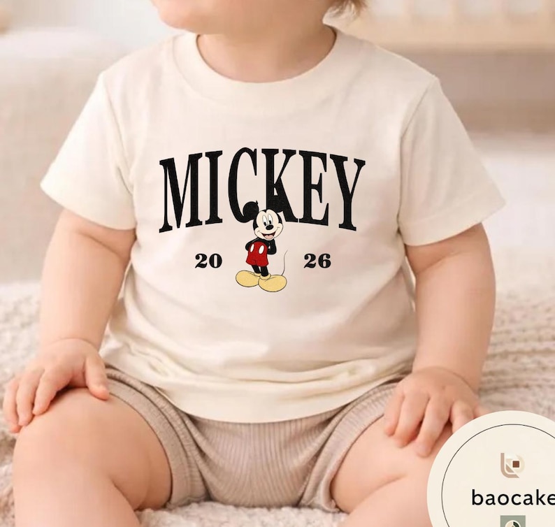May include: Cream-colored t-shirt featuring the word "MICKEY" in bold black letters. Below the word is a graphic of Mickey Mouse in his classic outfit, with the numbers "20" and "26" on either side. The shirt is short-sleeved.