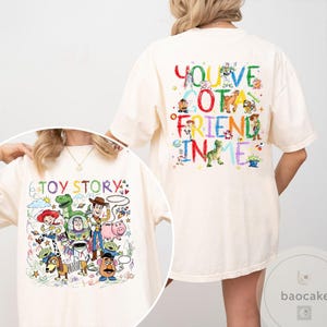 May include: Cream-colored t-shirt featuring colorful illustrations of characters from the animated film "Toy Story." The front of the shirt displays the text "Toy Story" with various characters. The back of the shirt has the phrase "You've Got a Friend in Me."