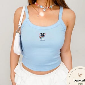 May include: A light blue ribbed tank top featuring a small embroidered graphic of the character Stitch wearing black mouse ears. The top has wide shoulder straps and a scoop neckline. A white shoulder bag is visible on the left side of the garment.