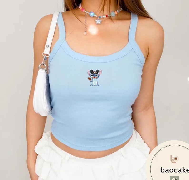 May include: A light blue ribbed tank top featuring a small embroidered graphic of the character Stitch wearing black mouse ears. The top has wide shoulder straps and a scoop neckline. A white shoulder bag is visible on the left side of the garment.