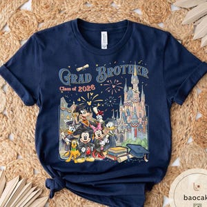 May include: Navy blue t-shirt with a colourful graphic featuring Disney characters, a castle, fireworks, and the text "Grad Brother Class of 2026". The design includes Mickey Mouse, Donald Duck, and Goofy.