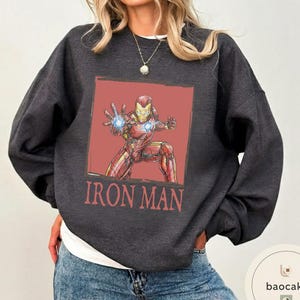 May include: Dark gray sweatshirt featuring a red and gold Iron Man graphic on a red background. The words "IRON MAN" are printed in red below the graphic. The sweatshirt is a crew neck style.