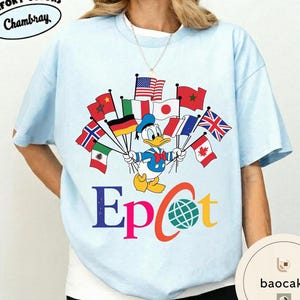 May include: Pale blue t-shirt featuring Donald Duck holding flags from different nations. The word "Epcot" is printed in colourful letters beneath the graphic. The shirt has the text "Comfort Colors Chambray" at the top.