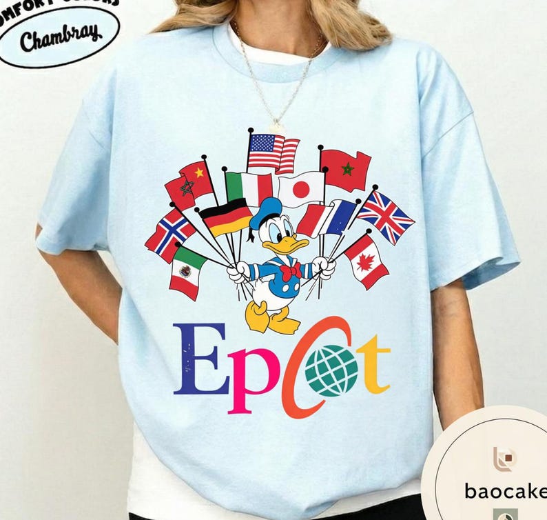 May include: Pale blue t-shirt featuring Donald Duck holding flags from different nations. The word "Epcot" is printed in colourful letters beneath the graphic. The shirt has the text "Comfort Colors Chambray" at the top.