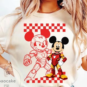 May include: Cream-colored t-shirt featuring a red and white checkered pattern and two Mickey Mouse illustrations. One Mickey is in a robotic suit, and the other is in an Iron Man suit. The shirt has a crew neck.