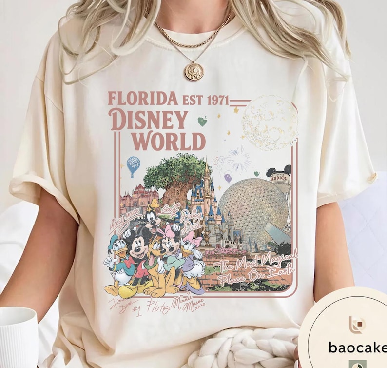 May include: A cream-colored t-shirt featuring a graphic print with the text FLORIDA EST 1971 DISNEY WORLD. The design includes illustrations of Disney characters, the Tree of Life, Epcot, and the text The Most Magical Place On Earth.