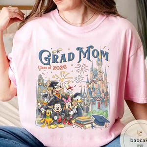 May include: Pale pink t-shirt with the words "Grad Mom" in blue script, and "Class of 2026" in smaller text. The graphic features Disney characters in graduation attire in front of a castle.