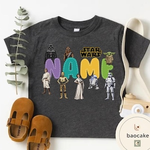 May include: Dark gray t-shirt featuring Star Wars characters and the word "NAME" in colorful letters. The shirt includes images of Darth Vader, Chewbacca, Yoda, and other iconic characters. Brown leather shoes are next to the shirt.
