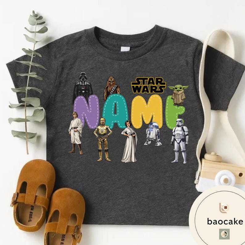May include: Dark gray t-shirt featuring Star Wars characters and the word "NAME" in colorful letters. The shirt includes images of Darth Vader, Chewbacca, Yoda, and other iconic characters. Brown leather shoes are next to the shirt.