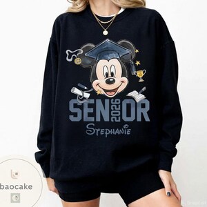 May include: Black sweatshirt featuring Mickey Mouse wearing a graduation cap. The text "SENIOR 2026" is displayed in gray, with the name "Stephanie" below. The design includes graduation-themed elements like a diploma and trophy.