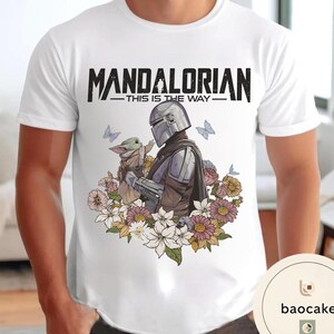 Puede incluir: Camiseta blanca con un dise&ntilde;o del Mandaloriano y Grogu rodeados de flores y mariposas. El texto "MANDALORIAN - THIS IS THE WAY -" est&aacute; impreso en negro en la parte superior.