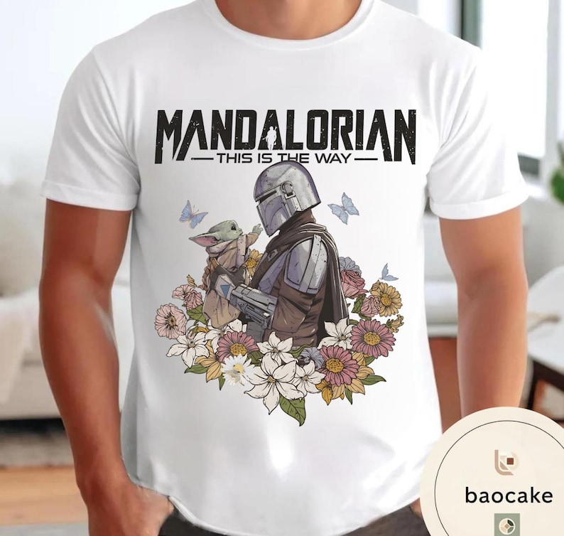 Puede incluir: Camiseta blanca con un dise&ntilde;o del Mandaloriano y Grogu rodeados de flores y mariposas. El texto "MANDALORIAN - THIS IS THE WAY -" est&aacute; impreso en negro en la parte superior.