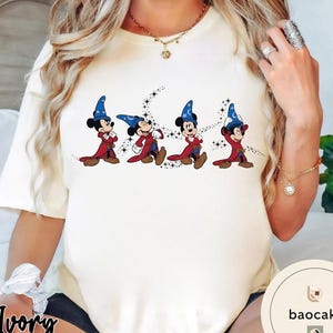 May include: Ivory-colored t-shirt featuring a graphic of Mickey Mouse in a wizard costume. The cartoon characters are wearing blue wizard hats and red robes. The design includes a starry background. The shirt is a crew neck.