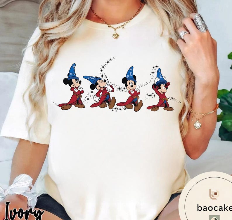 May include: Ivory-colored t-shirt featuring a graphic of Mickey Mouse in a wizard costume. The cartoon characters are wearing blue wizard hats and red robes. The design includes a starry background. The shirt is a crew neck.