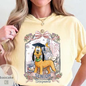 May include: Yellow t-shirt featuring a cartoon Pluto wearing a graduation cap, with the text "Senior Stephanie" and a Disney castle illustration. The design includes fireworks and floral accents.