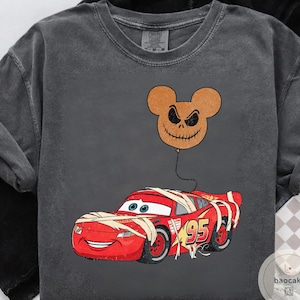 May include: Dark gray long-sleeve shirt featuring a red race car wrapped in bandages, with the number 95 on the side. A brown Jack Skellington-inspired balloon floats above the car. The shirt has a vintage, distressed look.