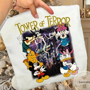 May include: White t-shirt featuring a graphic of Disney characters, including Mickey Mouse, Goofy, Minnie Mouse, Donald Duck, and Pluto, with the text "TOWER OF TERROR" in gold lettering. The background shows a building with lightning.