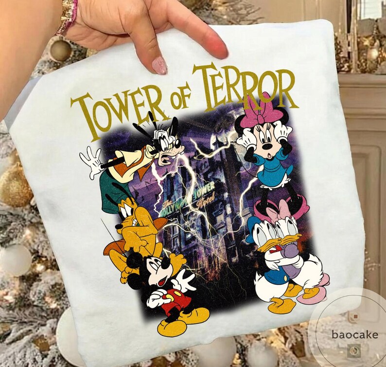 May include: White t-shirt featuring a graphic of Disney characters, including Mickey Mouse, Goofy, Minnie Mouse, Donald Duck, and Pluto, with the text "TOWER OF TERROR" in gold lettering. The background shows a building with lightning.