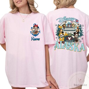 May include: Light pink t-shirt featuring a Minnie Mouse graphic with an anchor and the word "Name" in blue. The back of the shirt has a Disney Alaska 2026 design with various cartoon characters.