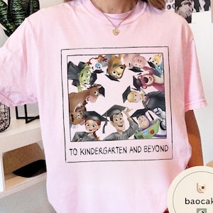 May include: Pale pink t-shirt with a framed graphic of animated characters wearing graduation caps. The graphic includes the text "TO KINDERGARTEN AND BEYOND". The shirt has a crew neck.