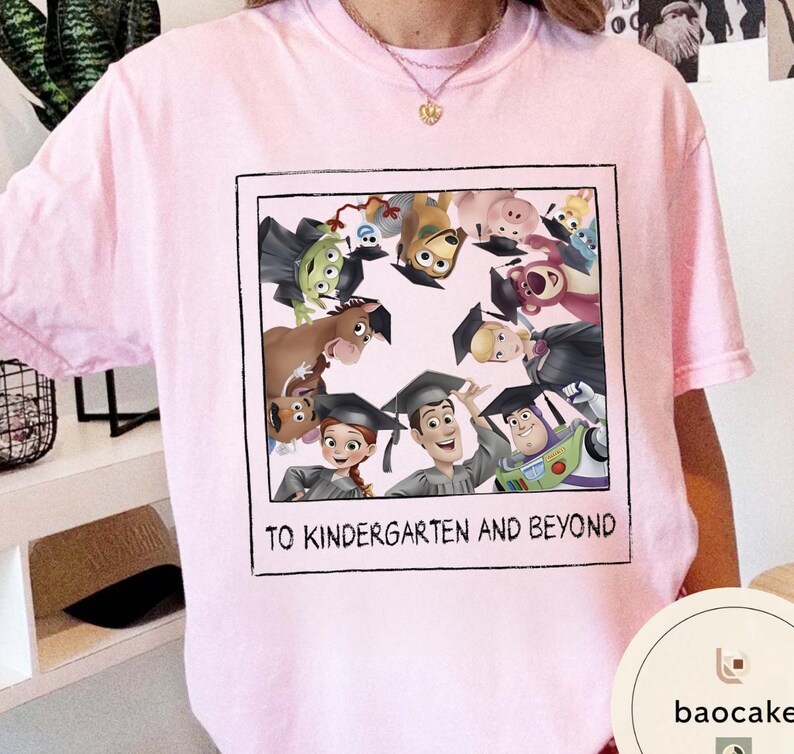 May include: Pale pink t-shirt with a framed graphic of animated characters wearing graduation caps. The graphic includes the text "TO KINDERGARTEN AND BEYOND". The shirt has a crew neck.