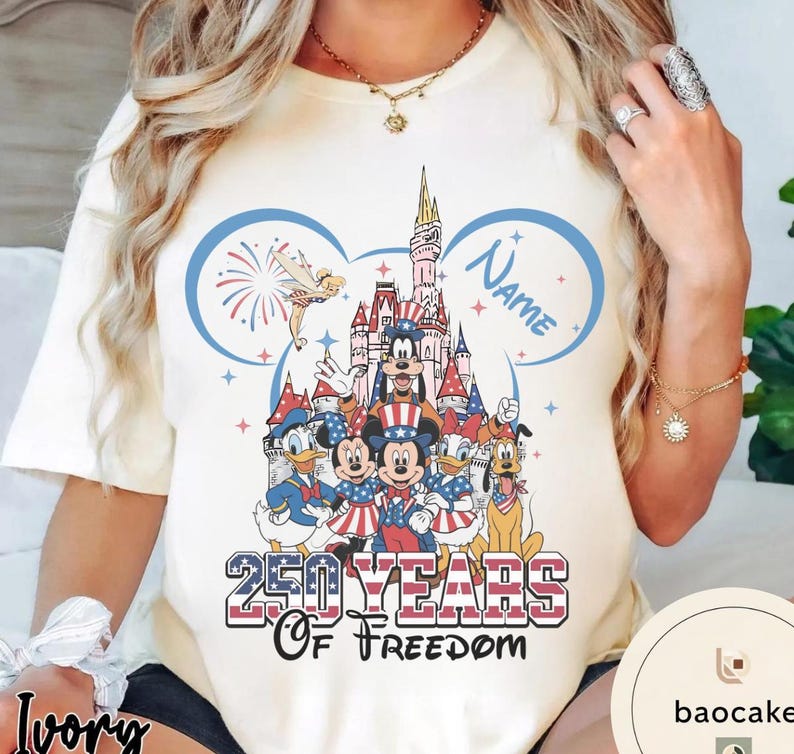 Peut inclure: T-shirt ivoire avec un motif patriotique repr&eacute;sentant Mickey Mouse et ses amis en rouge, blanc et bleu. Le motif comprend un ch&acirc;teau, des feux d'artifice et le texte "250 Years of Freedom". Un contour d'oreille de Mickey Mouse bleu encadre le motif.