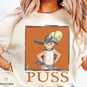 May include: Cream-colored sweatshirt featuring a cartoon illustration of a cat in a hat and cape, with the word "PUSS" in orange block letters. The cat is orange and white, with a black hat and cape.