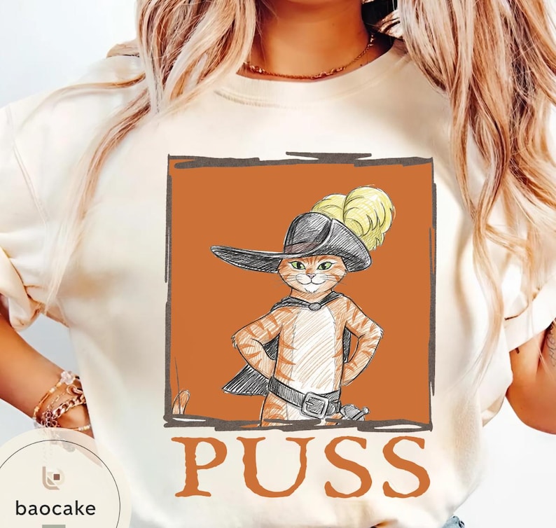 May include: Cream-colored sweatshirt featuring a cartoon illustration of a cat in a hat and cape, with the word "PUSS" in orange block letters. The cat is orange and white, with a black hat and cape.