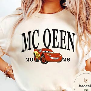 May include: Cream-colored sweatshirt featuring the text "MC QUEEN" in large black letters above a cartoon image of a red race car. The numbers "20" and "26" flank the car. The car has yellow and red accents.