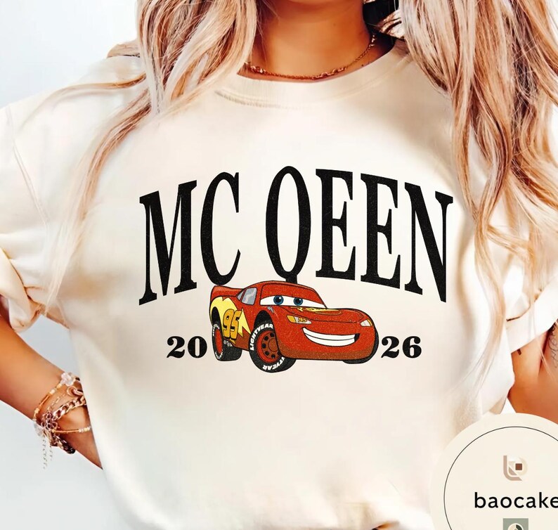 May include: Cream-colored sweatshirt featuring the text "MC QUEEN" in large black letters above a cartoon image of a red race car. The numbers "20" and "26" flank the car. The car has yellow and red accents.