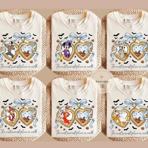May include: Cream-coloured t-shirts with Halloween designs. Each shirt features a heart-shaped locket with cartoon characters, bats, pumpkins, and the text "The most scariest place on earth".